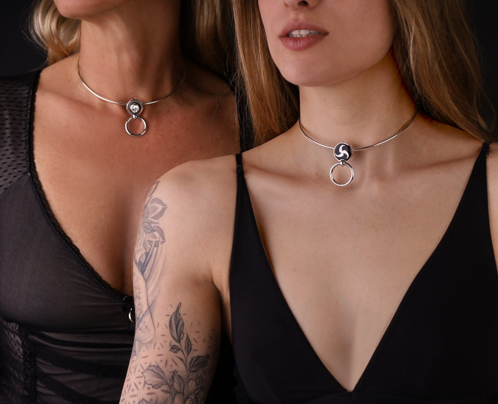 zwei Colliers an zwei Frauen. Das eine hat ein BDSM Peitschenrad, dass Zweite einen. Dir Ringe der O sind aufgeklappt. Das Collar ist aus Edelstahl